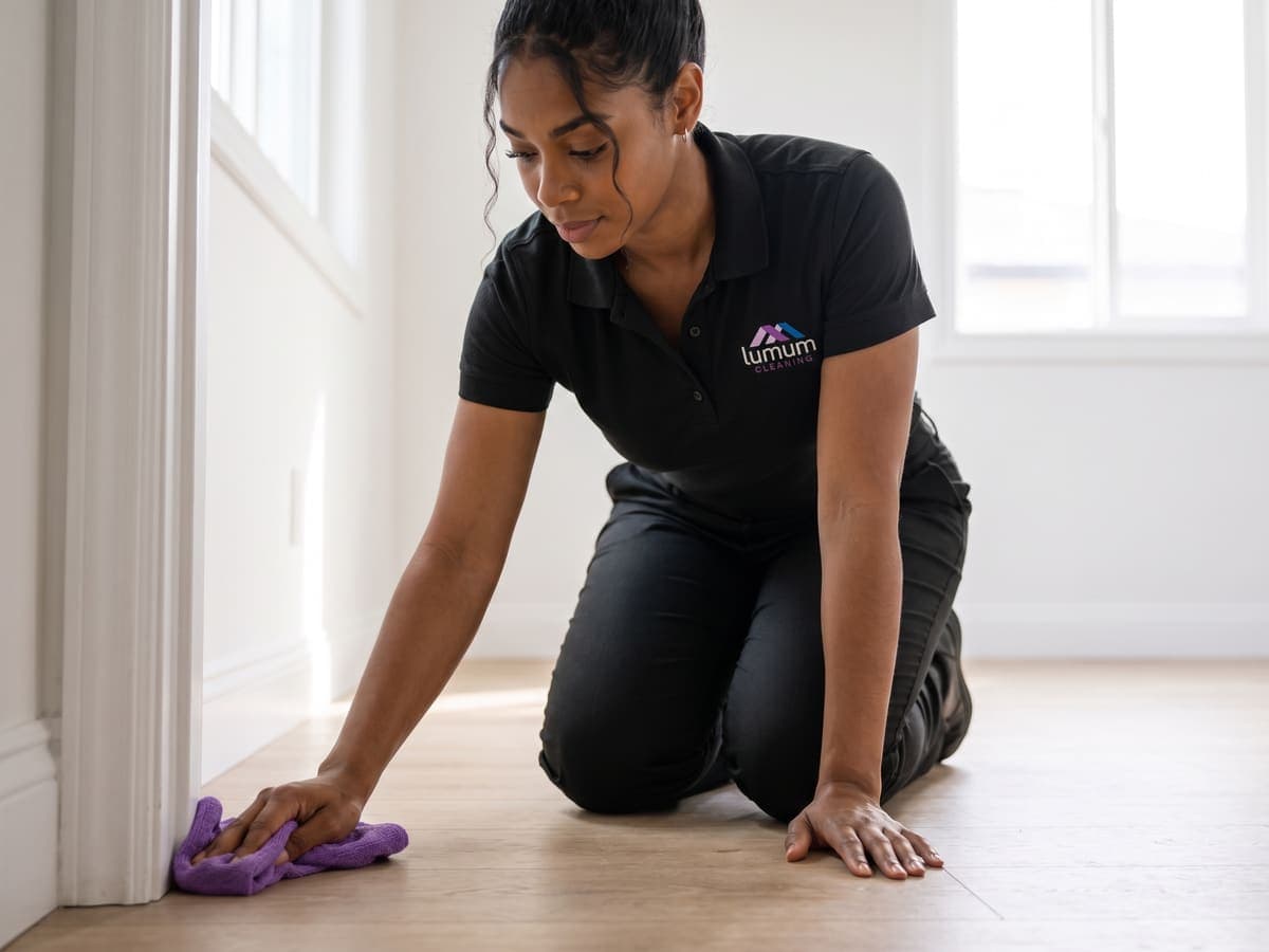 A Lumum Cleaning specialist wiping baseboards and trim with a purple microfiber cloth